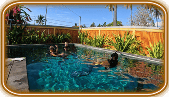 Tres buceadores en una piscina tropical rodeada de plantas y cerca de madera, practicando legend diving bajo luz solar.